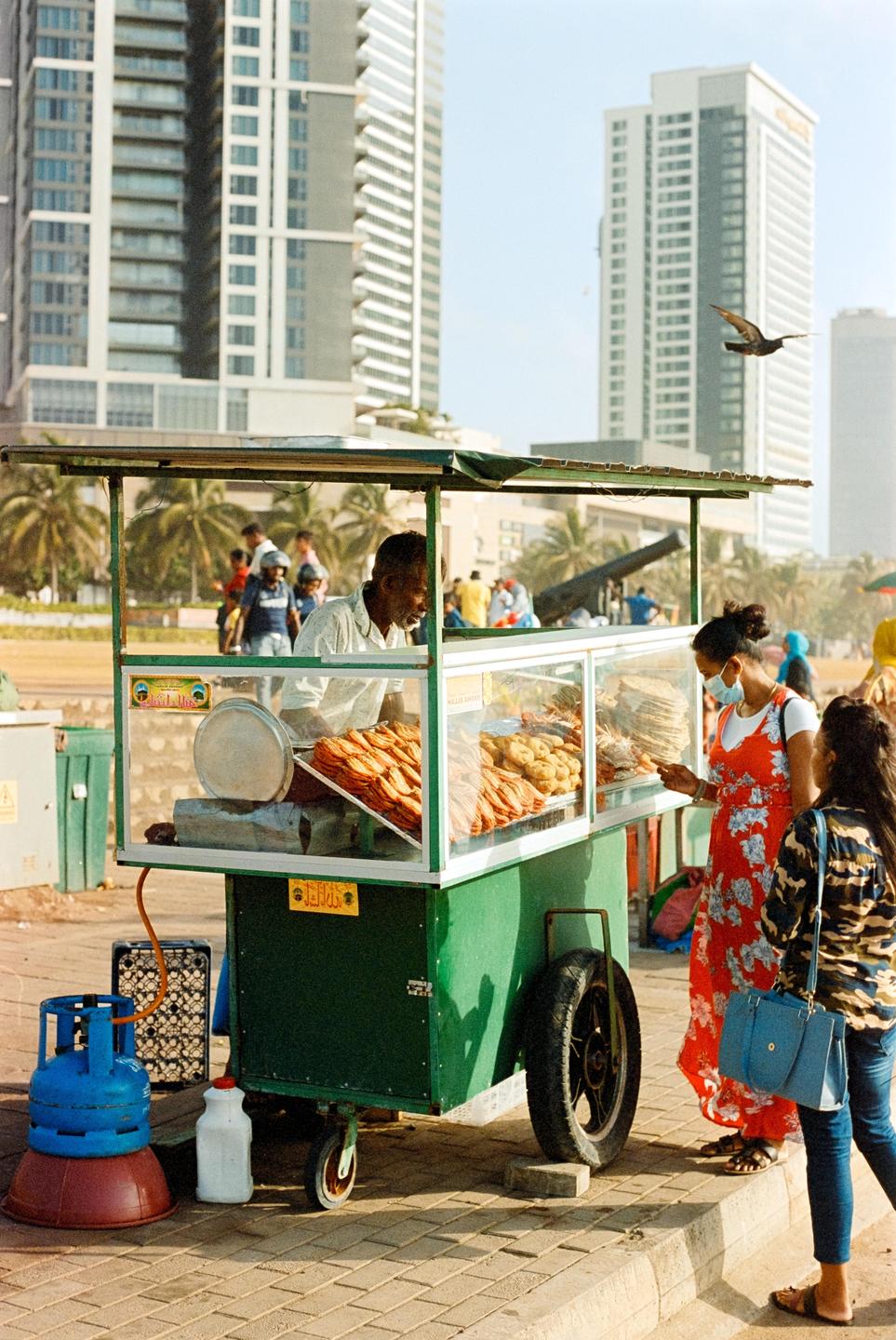 A man sells food from a street cart to two women on a sunny day in Colombo.