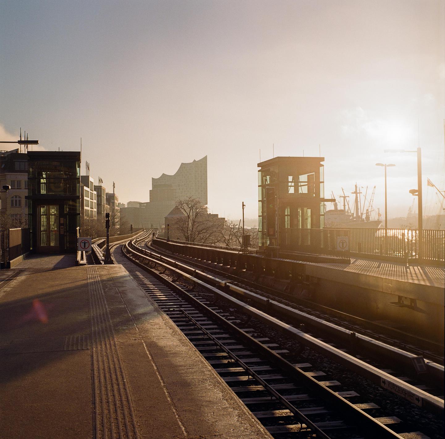 A train station platform and tracks curve towards a city skyline with a prominent modern building and a ship in the distance under a hazy sky.