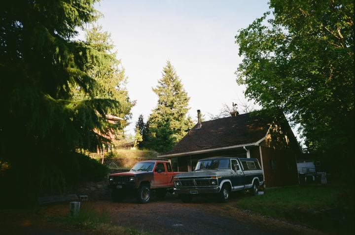 Two pickup trucks are parked on a dirt driveway next to a house surrounded by tall trees.