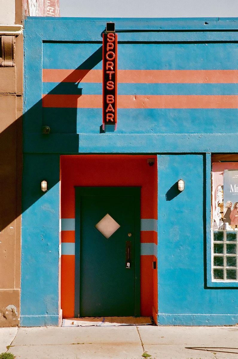 A sports bar with a blue and orange facade and a red neon sign is shown in bright sunlight.