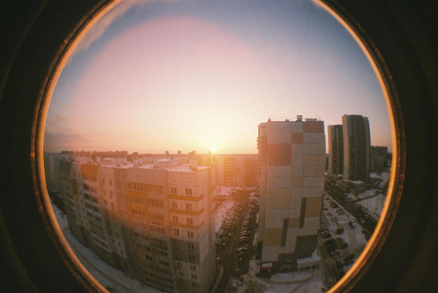 A fisheye view of a city at sunset shows apartment buildings and a parking lot covered in snow.