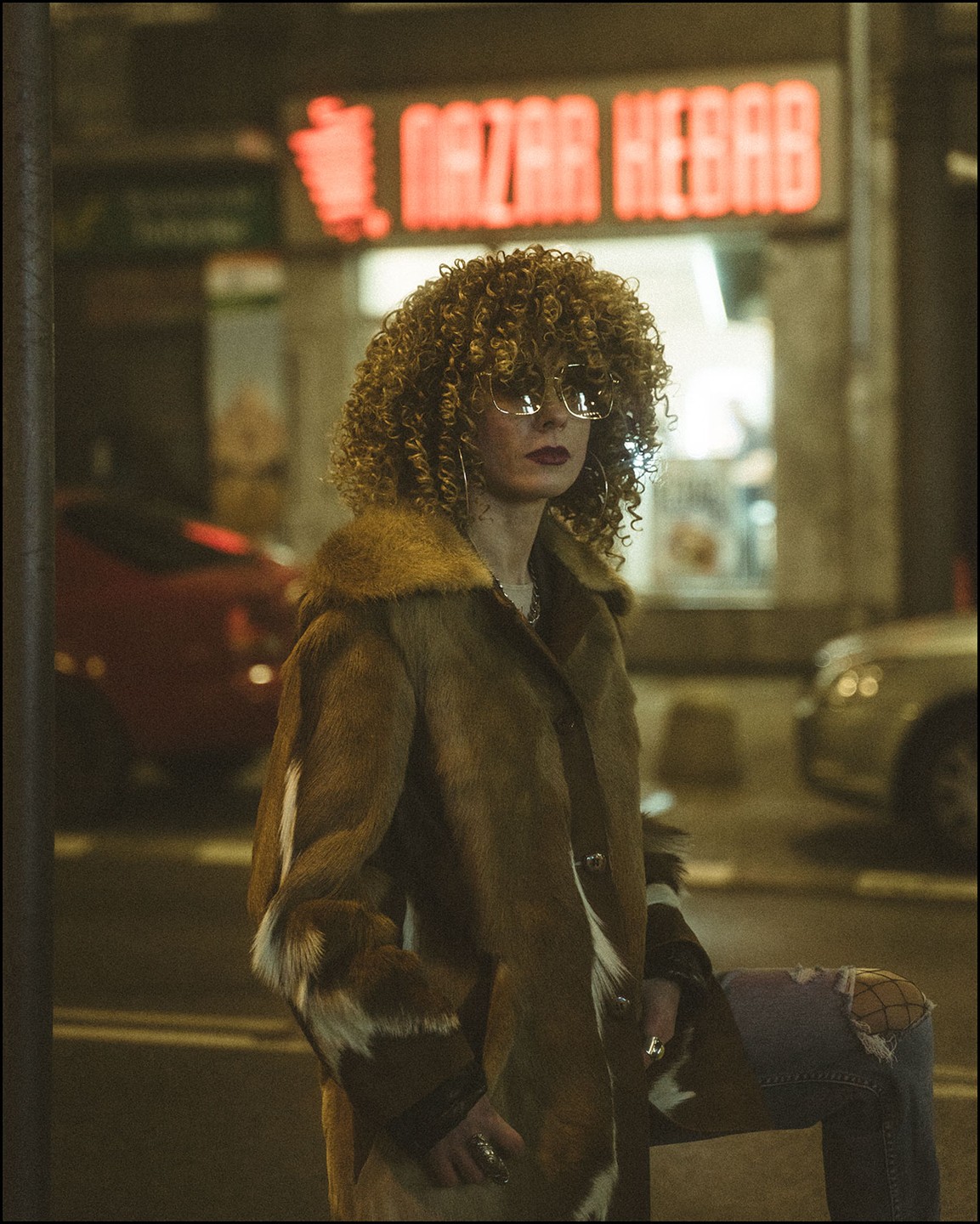 A woman with curly hair and sunglasses wears a fur coat and ripped jeans while standing on a street at night with a neon sign for Nazar Kebab in the background.