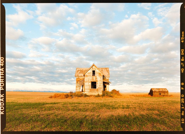 An abandoned house and a small shed stand in a vast field under a cloudy sky.