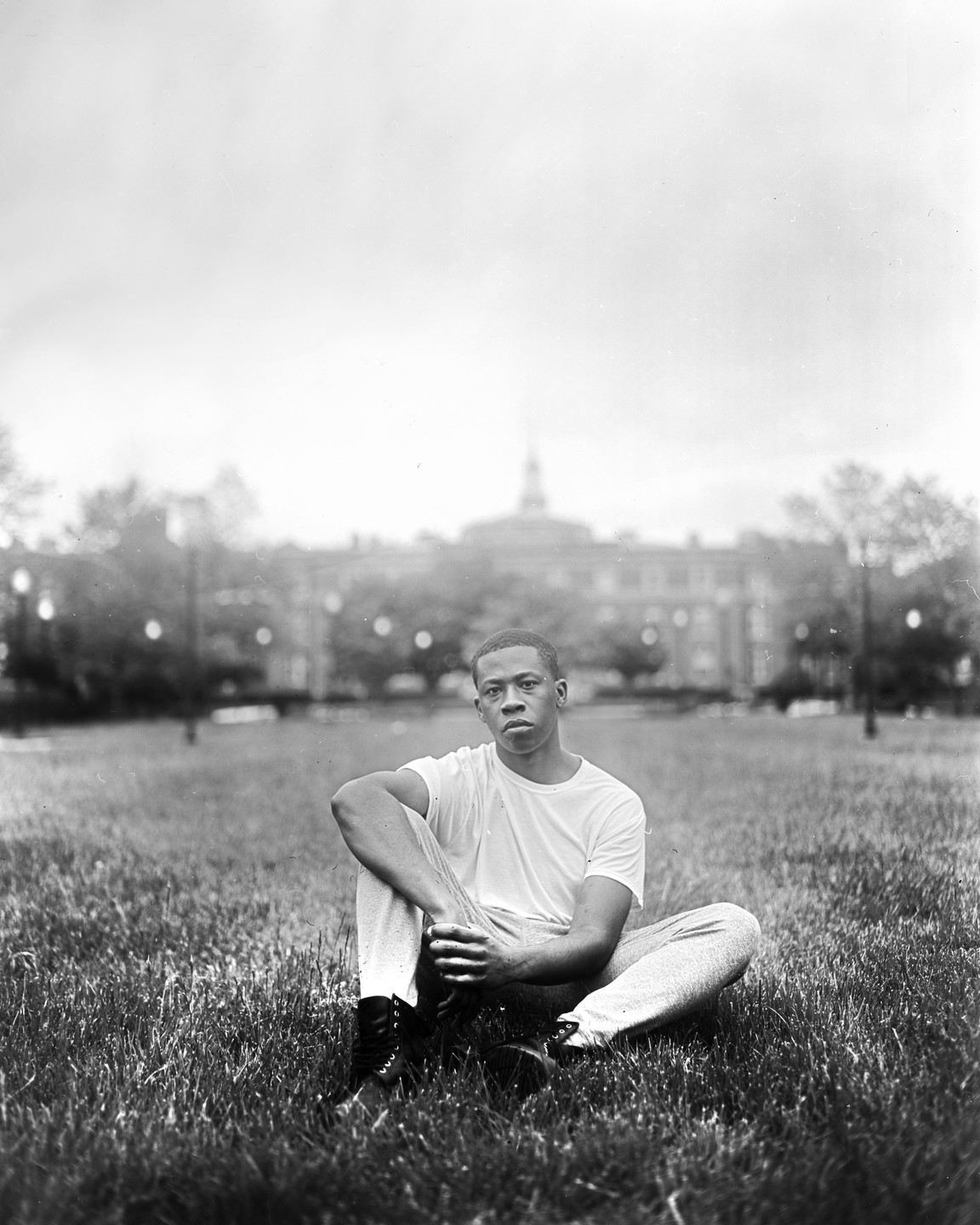 A man sits cross-legged in the grass with a building in the background.