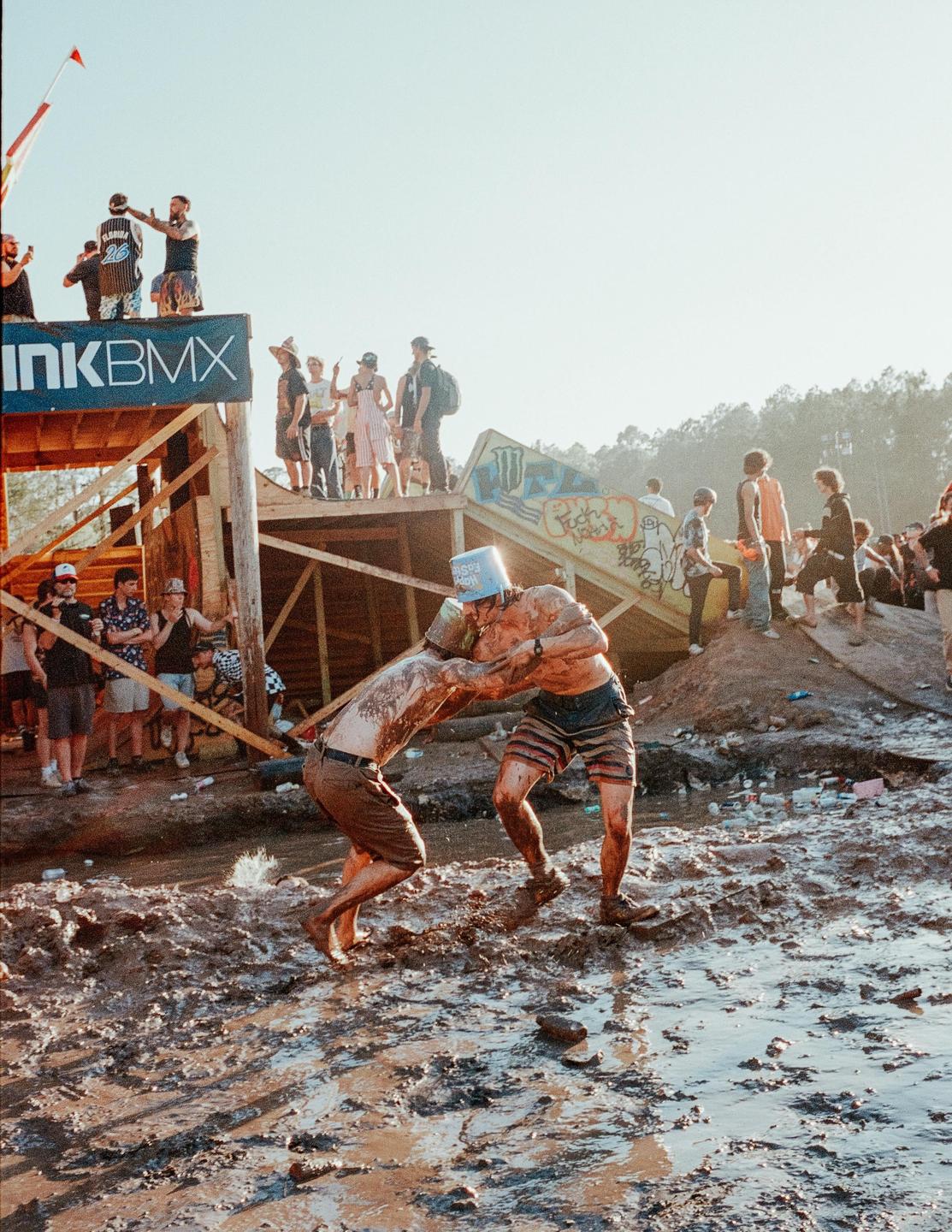 Two men covered in mud wrestle in a muddy pit while other people watch from a wooden structure.