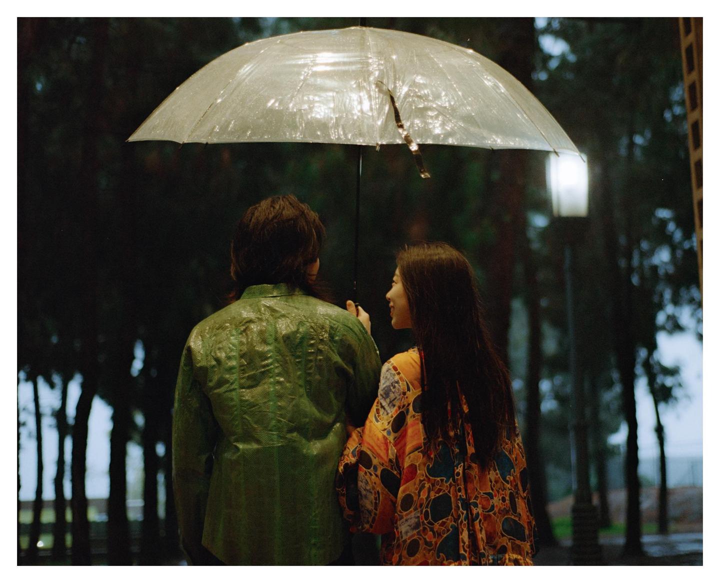 A couple stands under a clear umbrella in the rain at night.