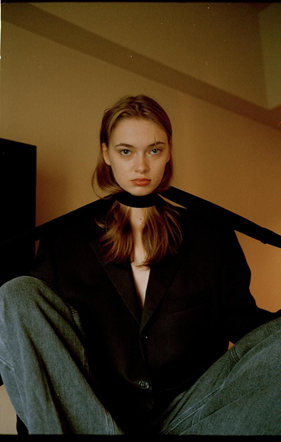 A woman wearing a black jacket and jeans sits with a black scarf around her neck.