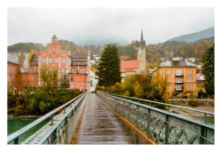 A wet bridge leads to a town with colorful buildings and a church steeple, surrounded by hills with autumn foliage.