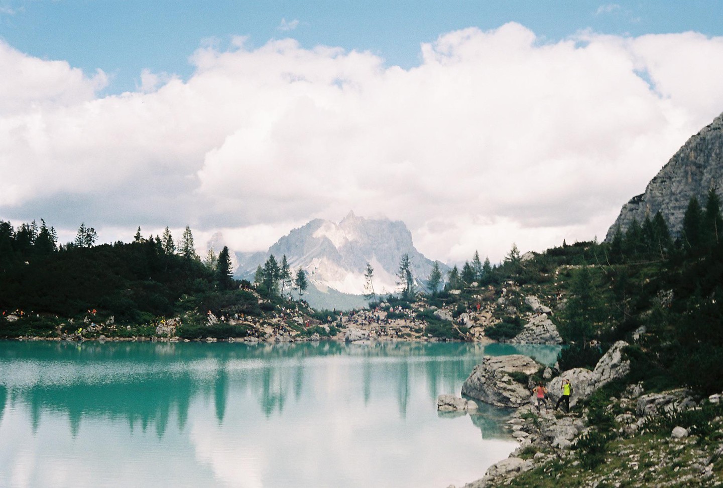 A large group of people are gathered around a turquoise lake with mountains and pine trees in the background.