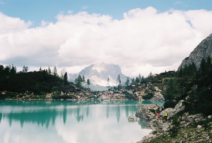 A large group of people are gathered around a turquoise lake with mountains and pine trees in the background.