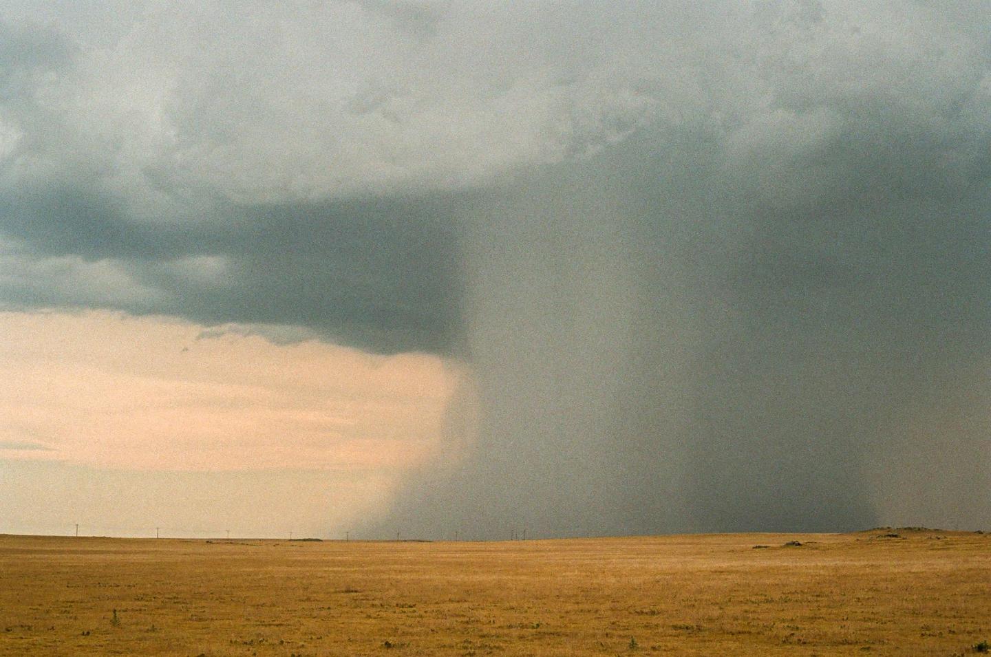 A large storm cloud with a visible rain shaft looms over a vast, flat, dry landscape.