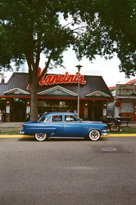 A blue vintage car is parked on the street in front of a restaurant with a large tree.