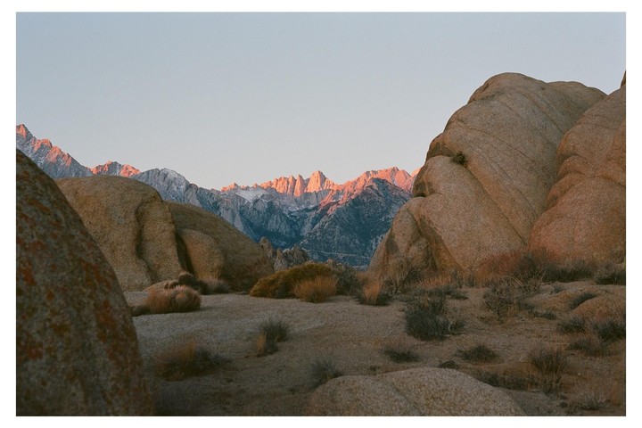 A desert landscape features large rock formations in the foreground and snow-capped mountains in the distance, illuminated by the golden hour light.
