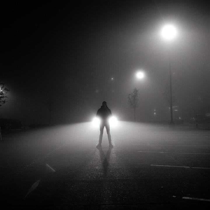 A man stands in front of a car with its headlights on in a foggy parking lot at night.