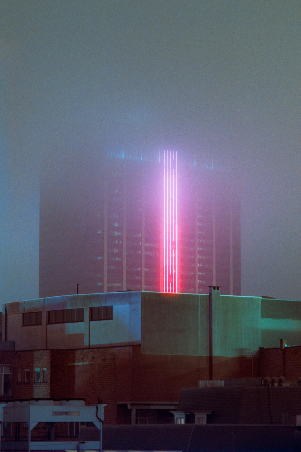 A tall building with a bright pink light beam is partially obscured by fog, with other buildings visible in the foreground.