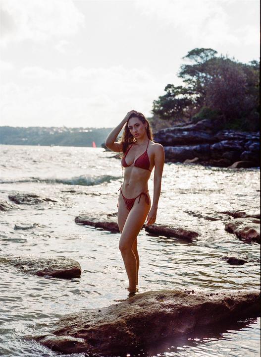 A woman in a red bikini stands in shallow water among rocks with a body of water and distant land in the background.