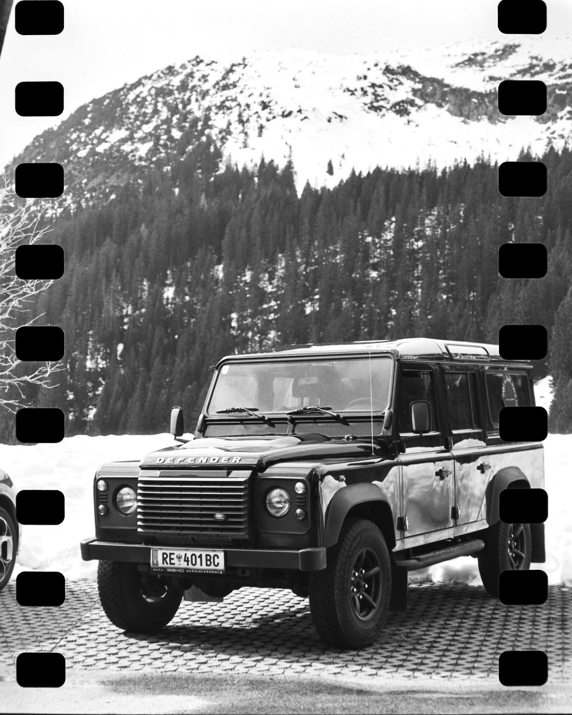 A black Land Rover Defender is parked on a paved surface with a snow-covered mountain and forest in the background.