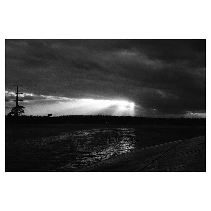Sunlight breaks through dark clouds over a body of water with a distant tree line and a power pole.