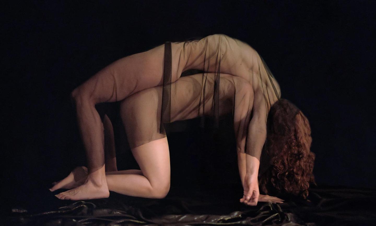 A woman in a sheer black garment is posed in a backbend on a dark surface.