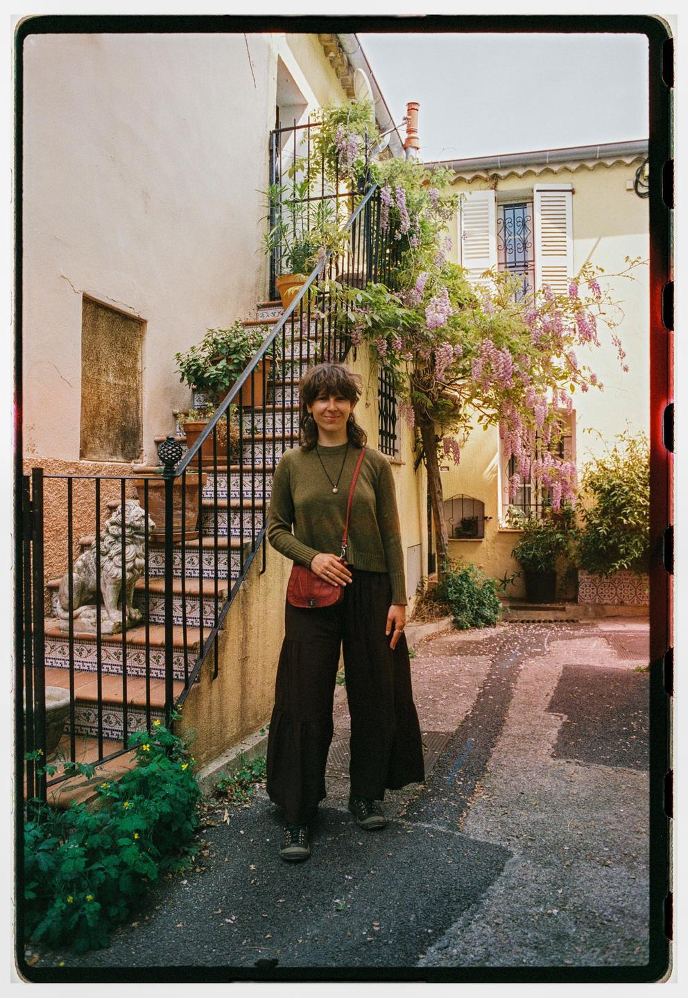 A woman stands on a paved path in front of a building with an outdoor staircase adorned with flowering vines.