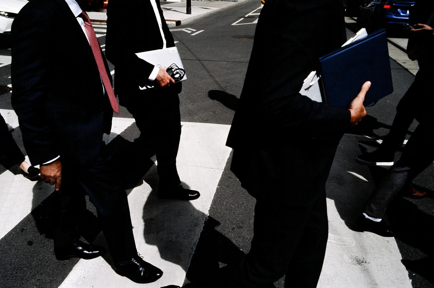 Several men in suits walk across a crosswalk, casting long shadows on the street.