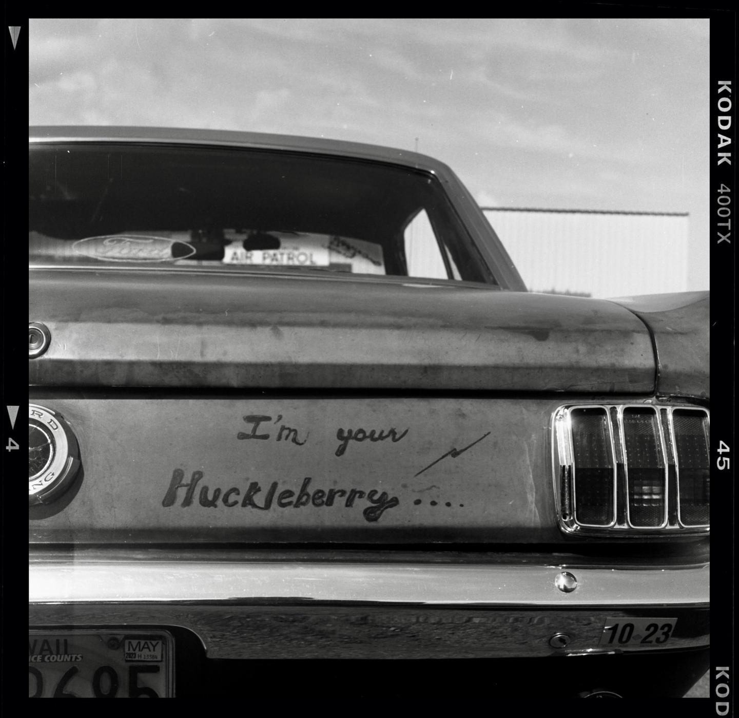 The rear of a vintage car with the phrase "I'm your Huckleberry" written on the trunk is visible.