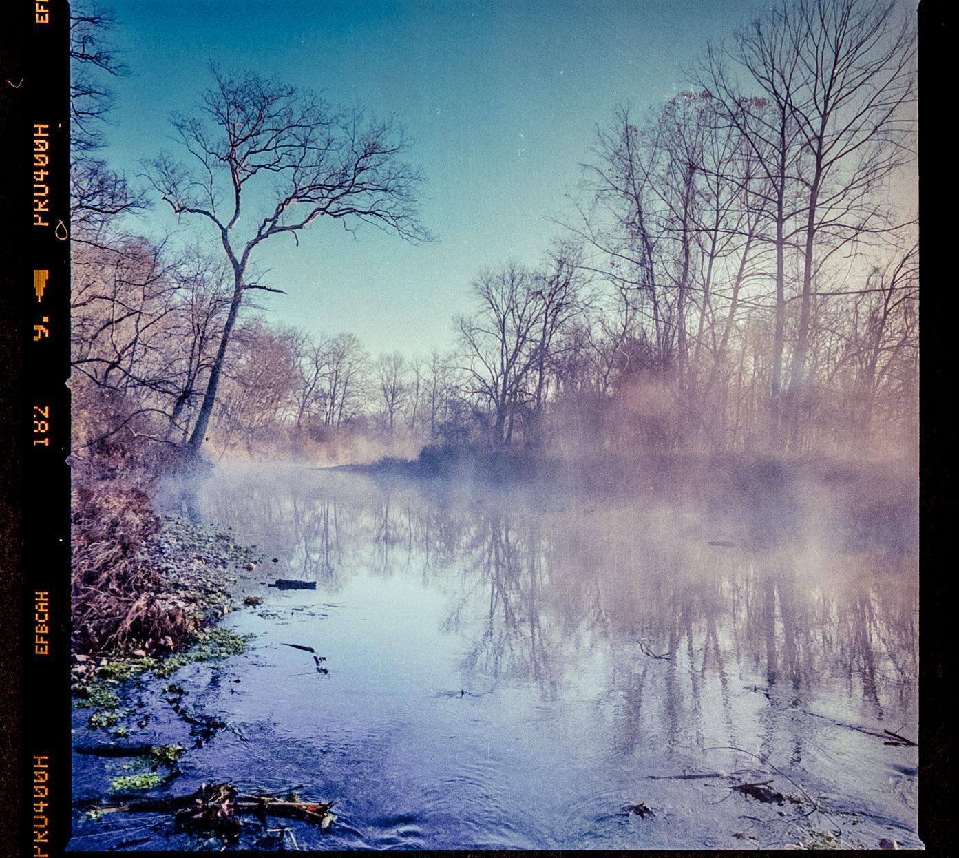 A misty river with bare trees on its banks is shown under a clear sky.