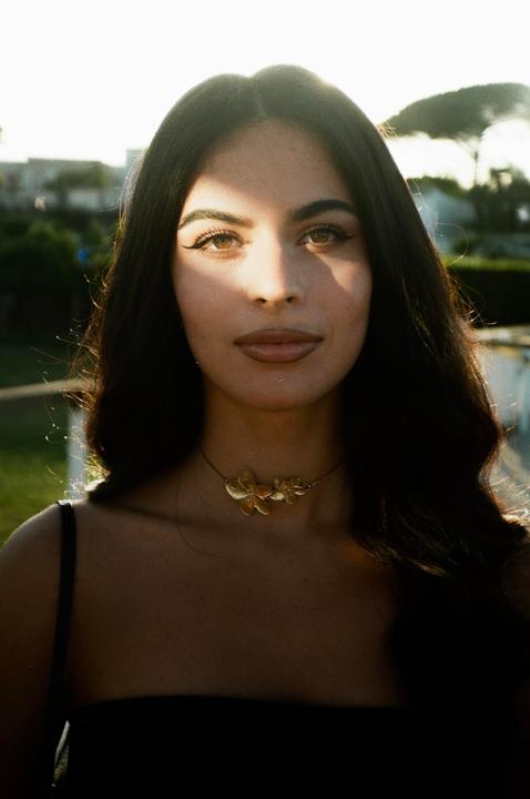 A woman with long dark hair and a gold floral necklace looks directly at the viewer.