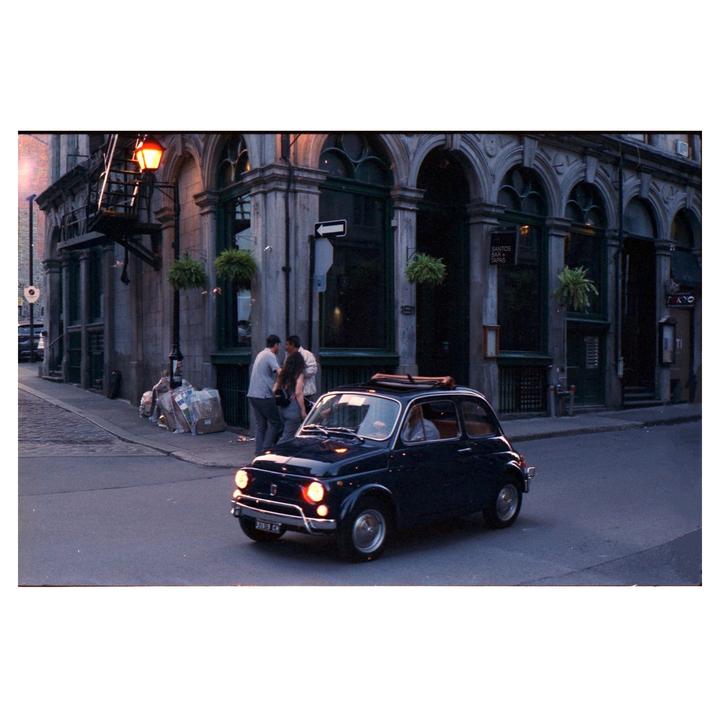 A small dark car with its headlights on drives past a building with arched windows and a street light.