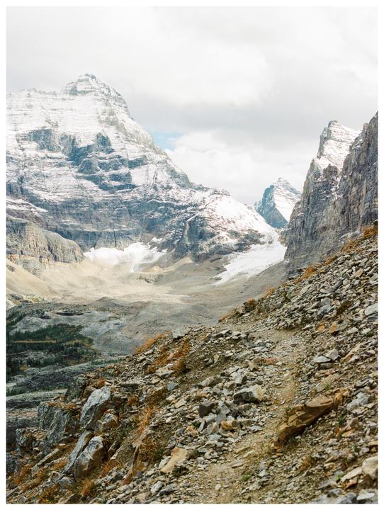 A rocky mountain path leads down into a valley with snow-capped mountains and a glacier.