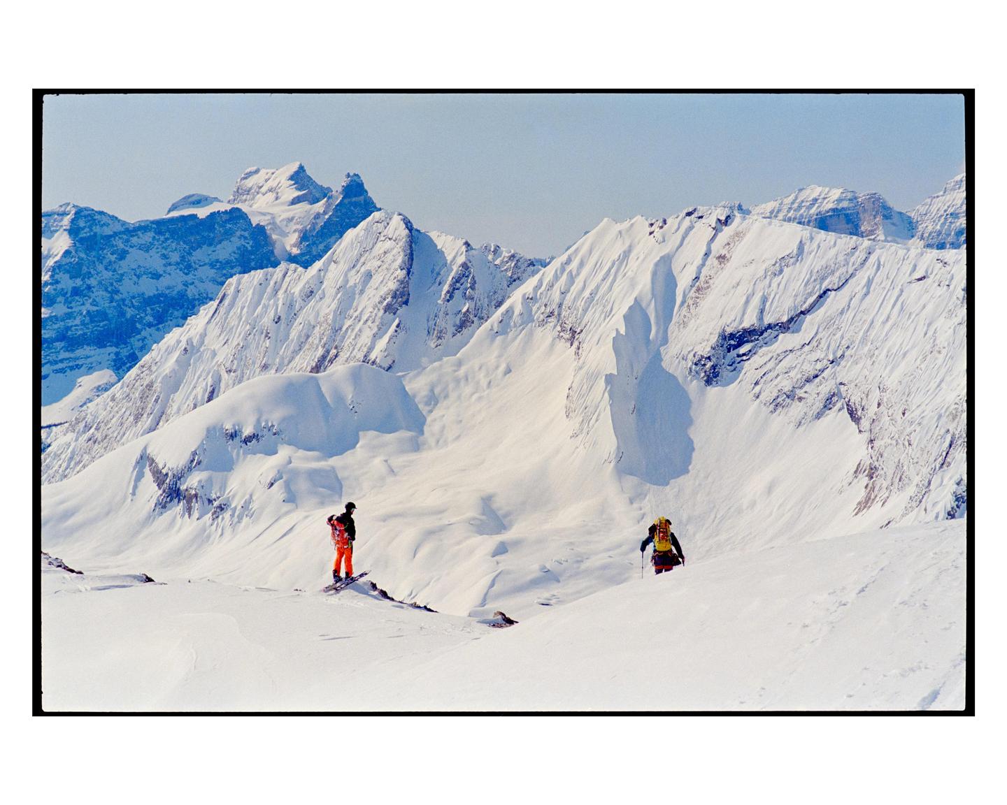 Two people are in a snowy mountain landscape, one on a snowboard and the other walking with poles.
