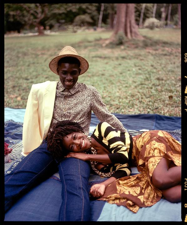A man in a straw hat sits on a blanket with a woman resting her head on his lap in a grassy area.