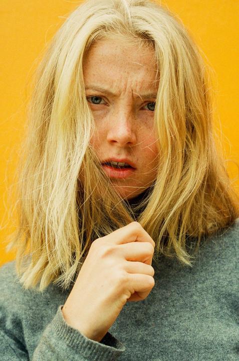 A woman with blonde hair and freckles looks directly at the viewer with a furrowed brow against a yellow background.