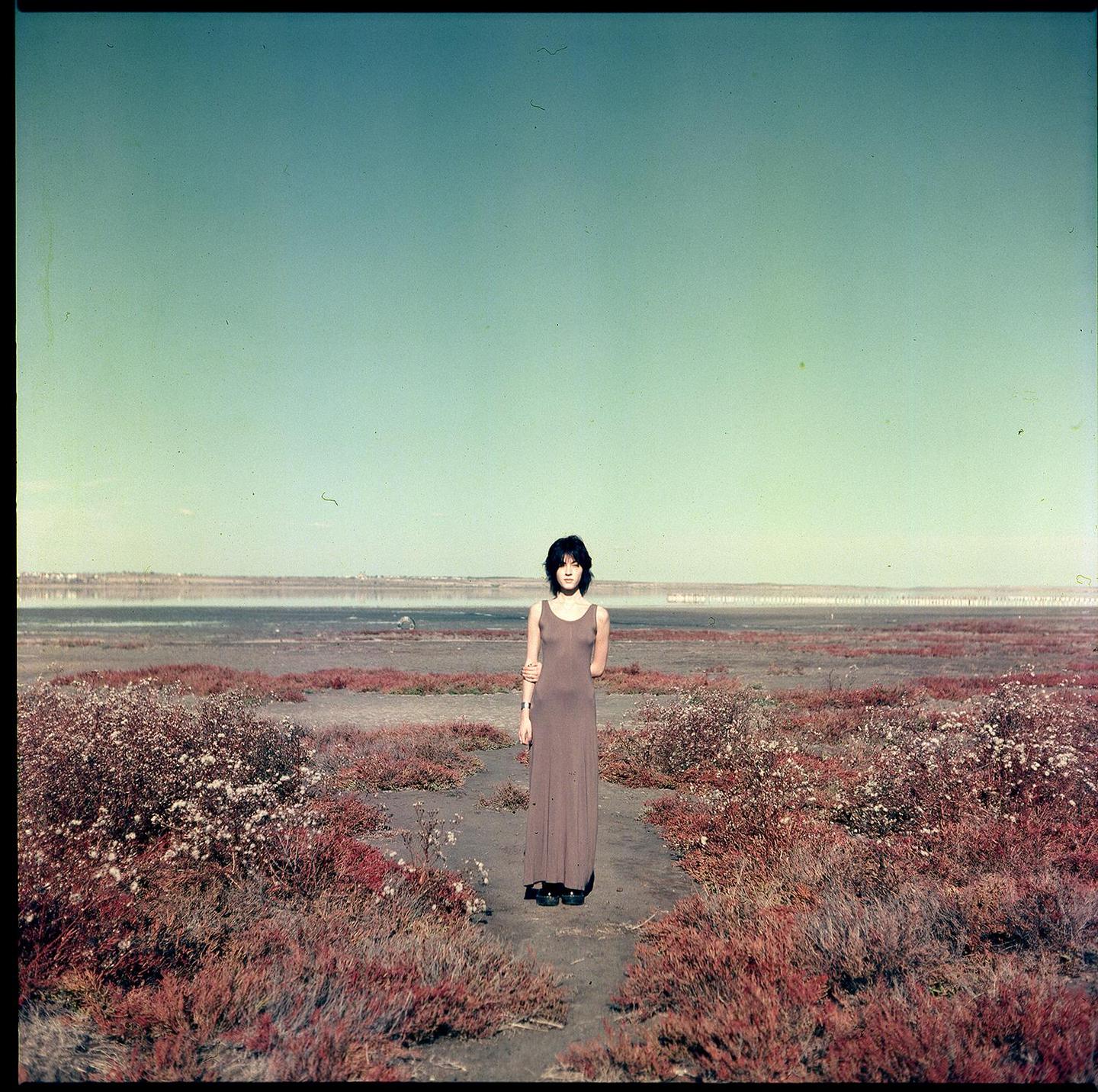 A woman stands in a barren landscape with red bushes and a body of water in the distance.