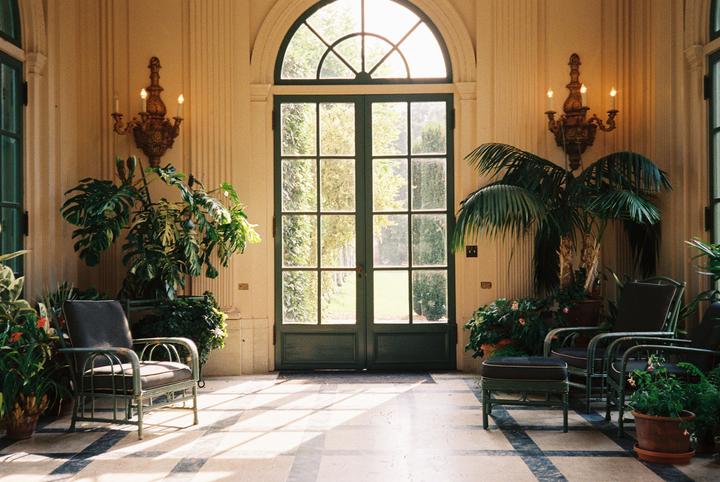 Sunlight streams through a large arched glass door into a room with potted plants and chairs.