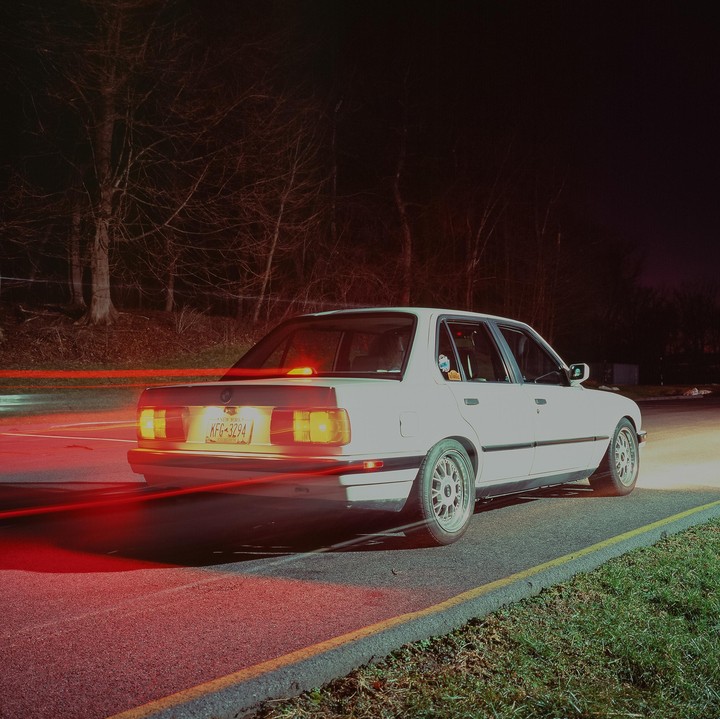 A white car is parked on the side of a road at night, with red light trails streaking past its rear.