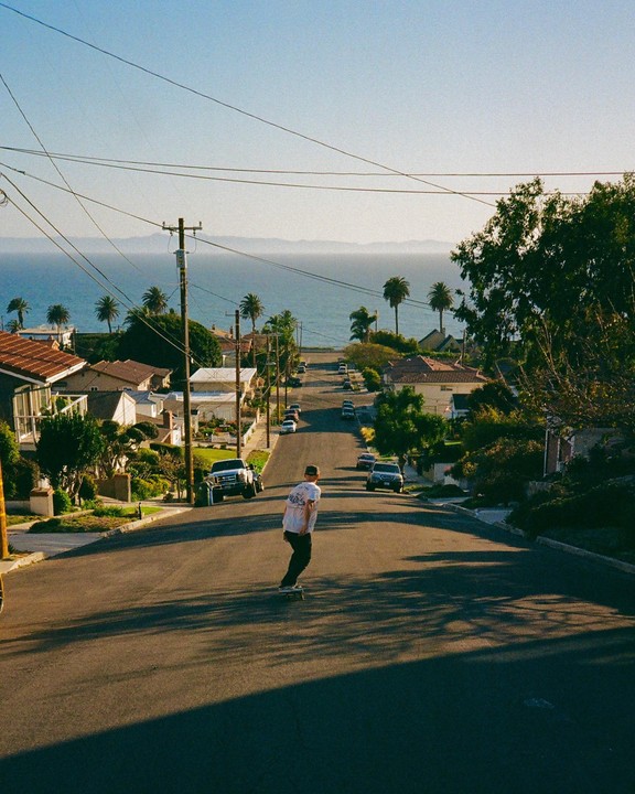 A man on a skateboard rides down a residential street towards the ocean.