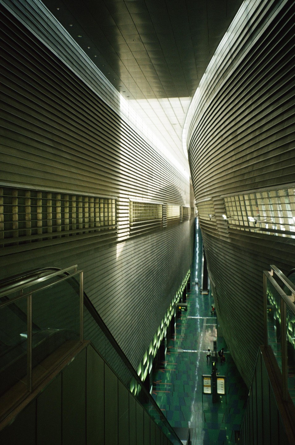 A long, narrow, modern subway station with a green floor and escalators on either side is illuminated by natural light from above.