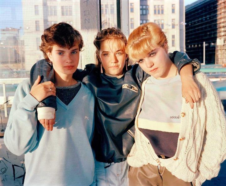 Three young women stand together with their arms around each other in front of a window with buildings visible outside.