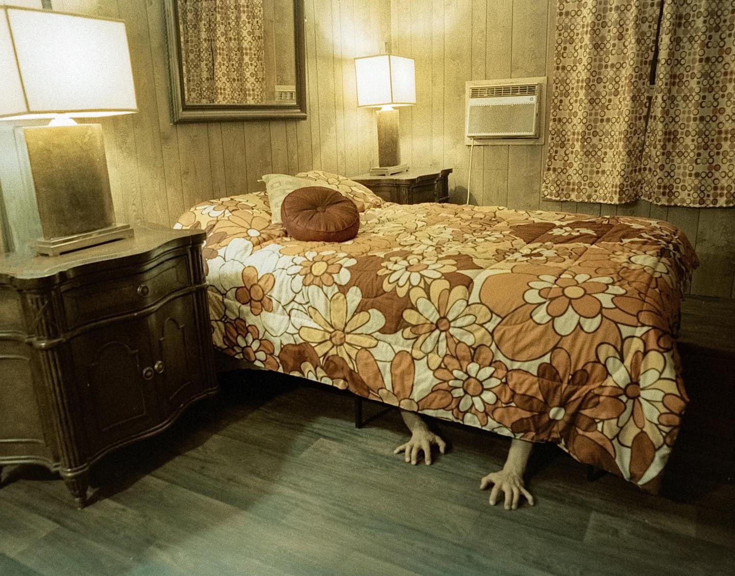 A pair of hands with splayed fingers reaches out from under a bed with a floral comforter in a dimly lit room.