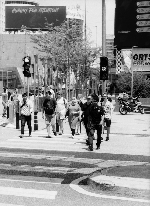 A group of people crosses a street at a crosswalk with a billboard in the background that reads "Hungry for Attention?".