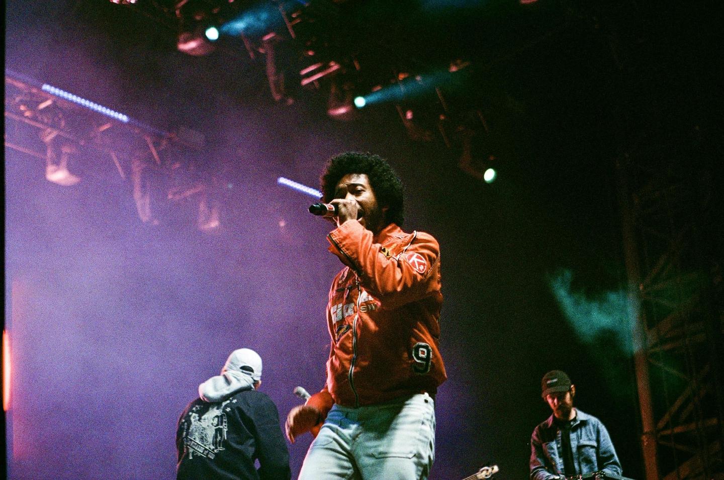 A man with an afro sings into a microphone on stage, illuminated by purple and green stage lights.