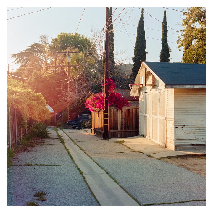 A narrow alleyway leads past a white garage with a basketball hoop and a fence covered in pink bougainvillea.