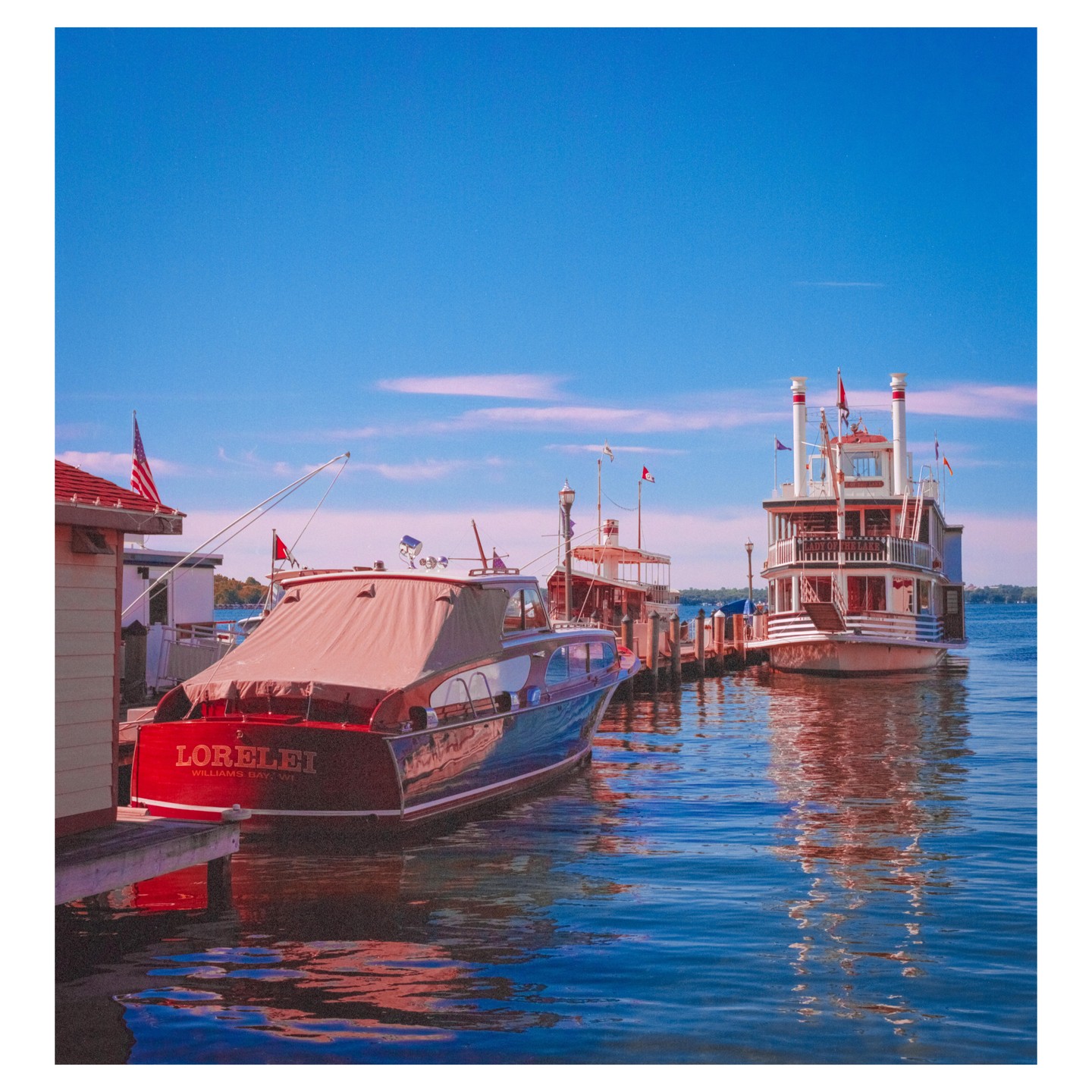 A red and blue boat is docked next to a large white paddle boat on a clear day.