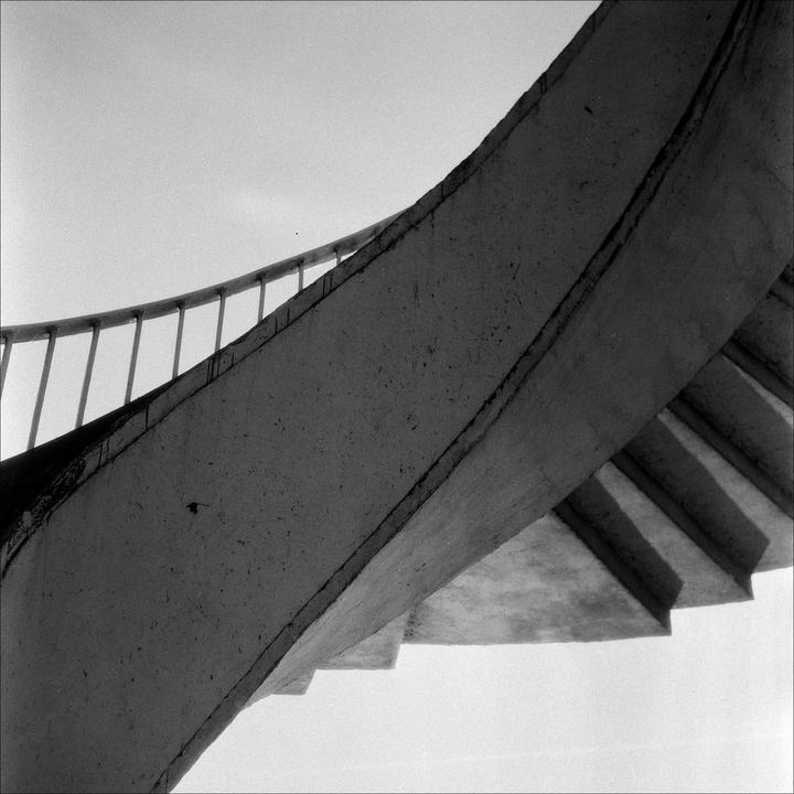 A concrete spiral staircase with a metal railing ascends towards a bright sky, casting shadows on its underside.