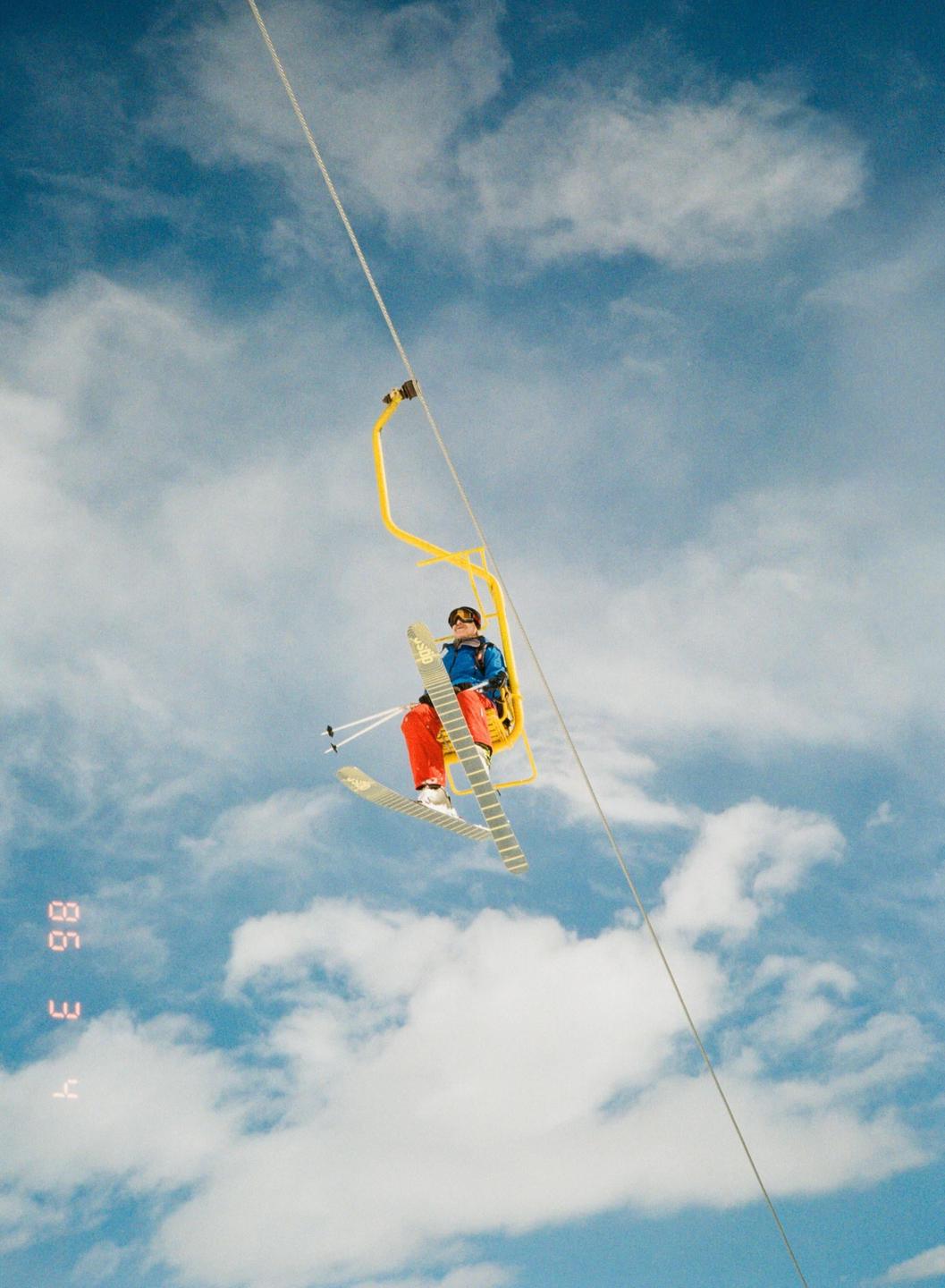 A person in ski gear is riding a yellow ski lift against a cloudy blue sky.