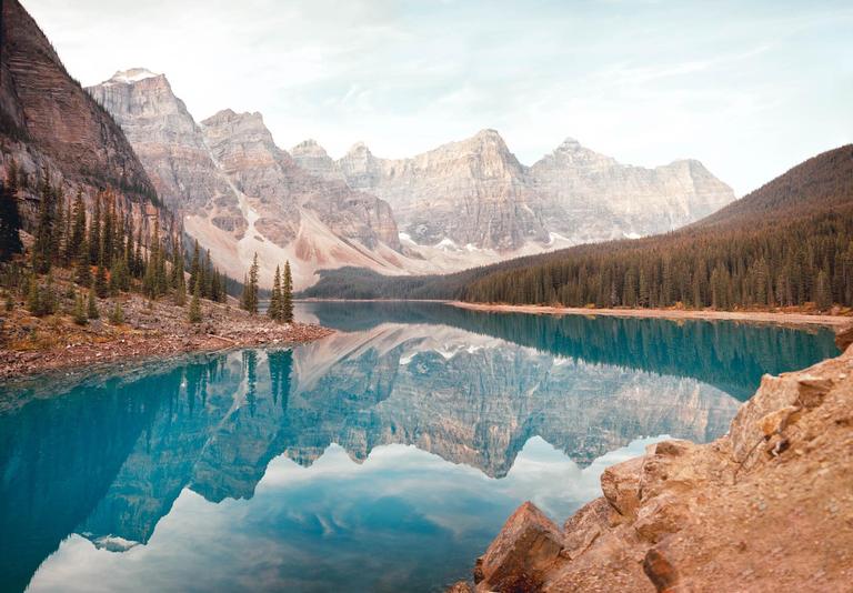 A panoramic landscape shows a turquoise lake reflecting mountains and a forest under a hazy sky.