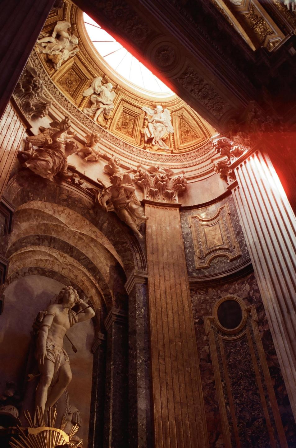 A grand interior with ornate sculptures and columns is illuminated by light streaming through a high window.