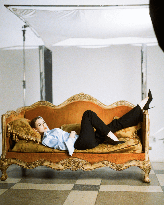 A woman in a light blue shirt and black pants is lying on an ornate orange sofa with her legs up.