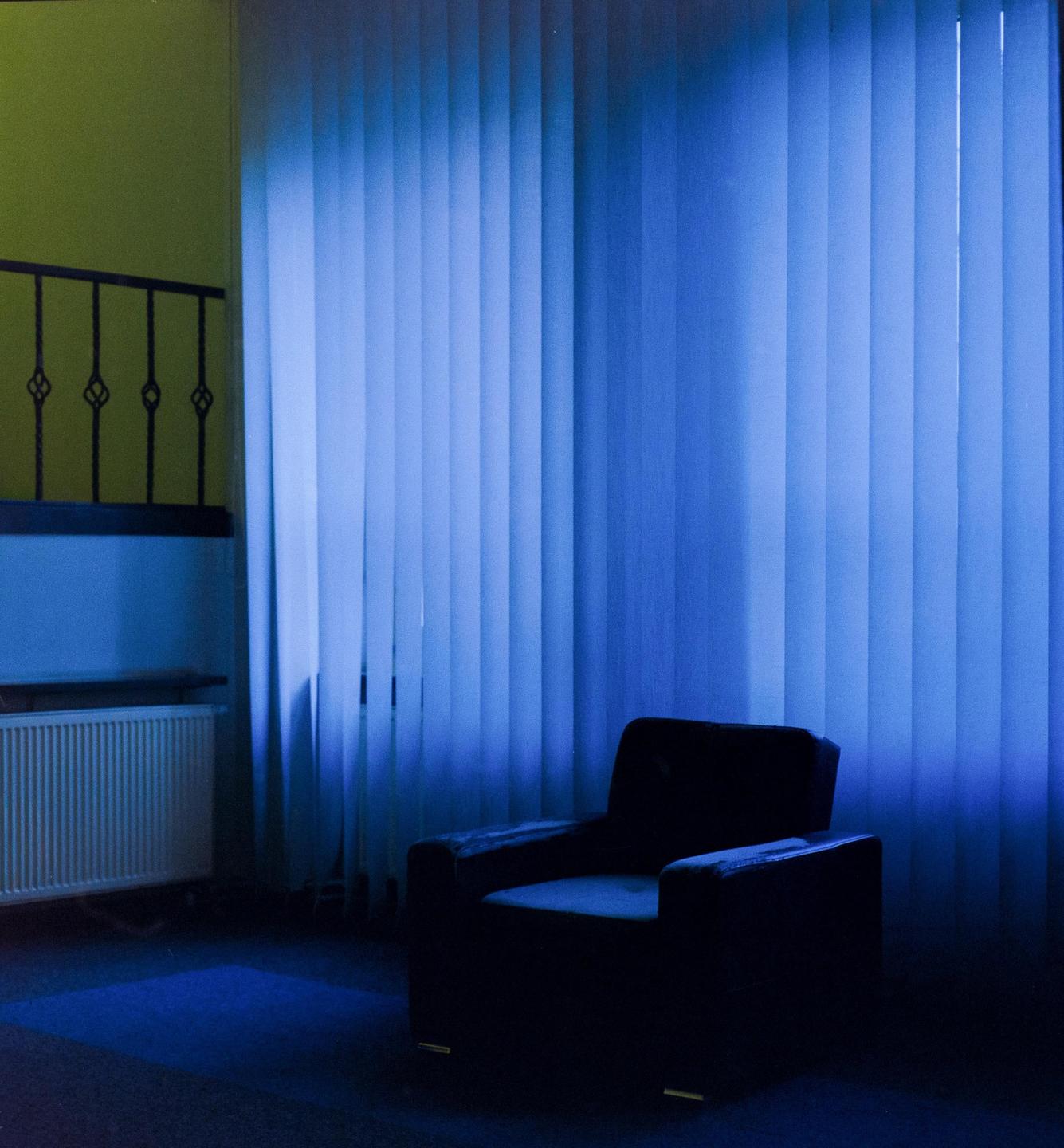 An armchair sits in a room with blue light coming through vertical blinds.